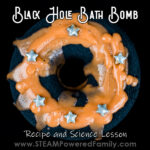 A black donut shaped bath bomb sits on a black background. The bath bomb is covered with a ring of orange soap, with a sprinkling of silver stars. Overlay text says Black Hole Bath Bomb Recipe and Science Lesson