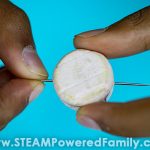 A child's fingers show how to insert the needle into a cut off wine cork while making a homemade compass