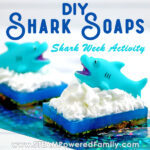 On a white background with a glittery turquoise foil strip sits two soaps with a dark base, blue middle, and white frothy waves with a shark cresting out of the waves. Overlay text says DIY Shark Soaps Shark Week Activity