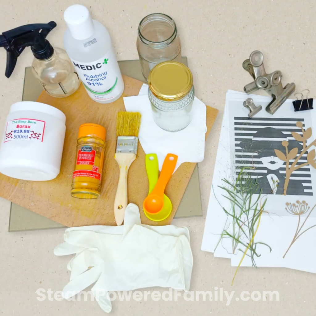 Materials for sun printing with turmeric arranged on a wooden board, including turmeric powder, borax, rubbing alcohol, a paintbrush, measuring spoons, rubber gloves, a spray bottle, and stencils with pressed grasses.
