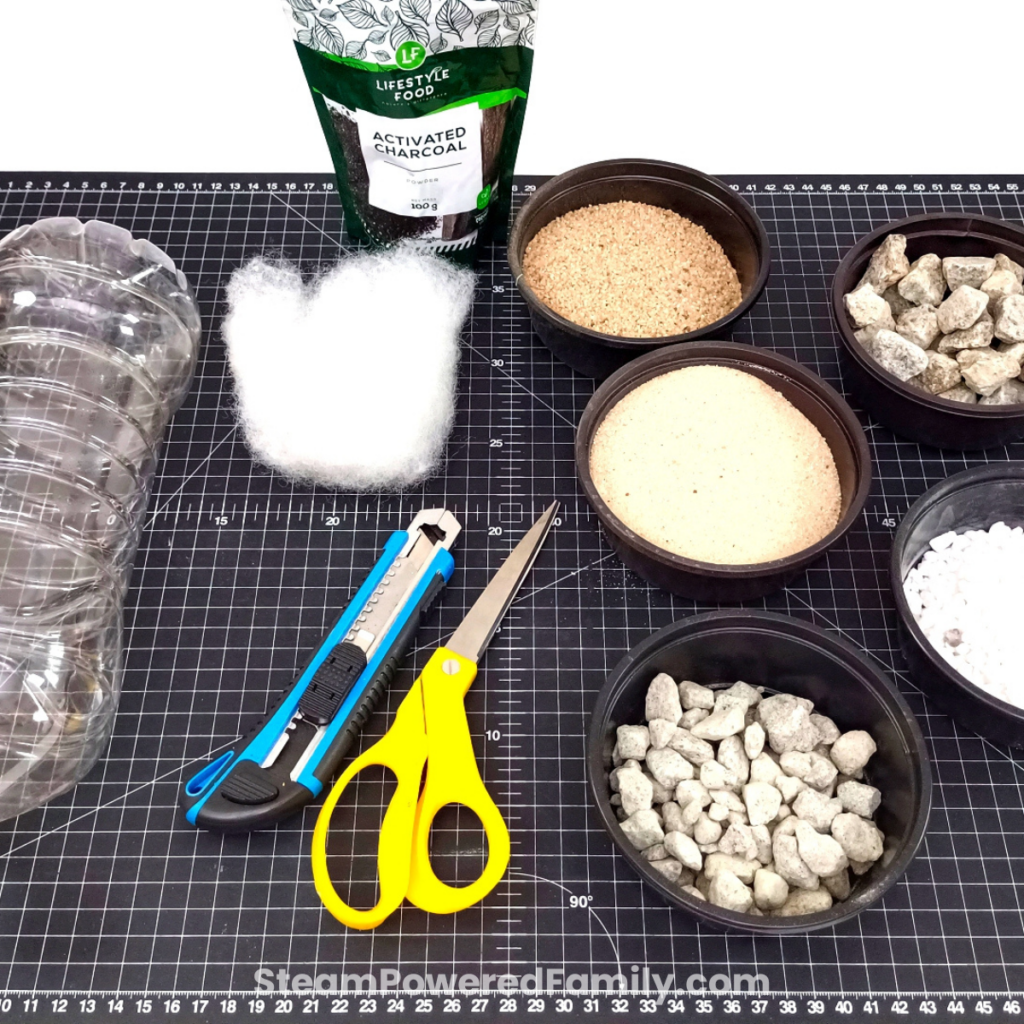 All the materials for a water filtration experiment laid out, including a plastic bottle, activated charcoal, sand, stones, and polyester batting.